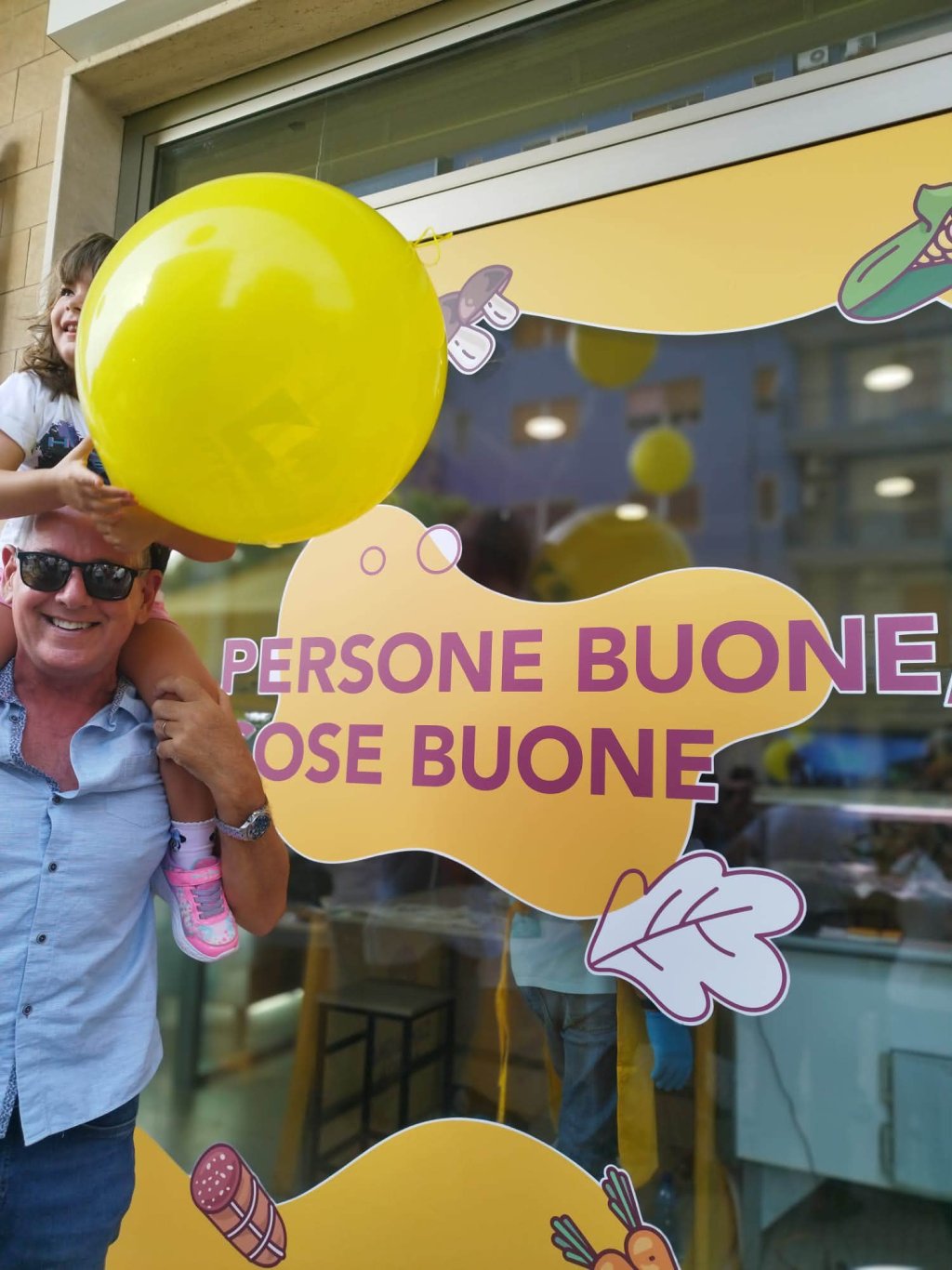 Uomo porta bambina sulle spalle, palloncino giallo, cartellone con scritta \Persone buone, cose buone\
