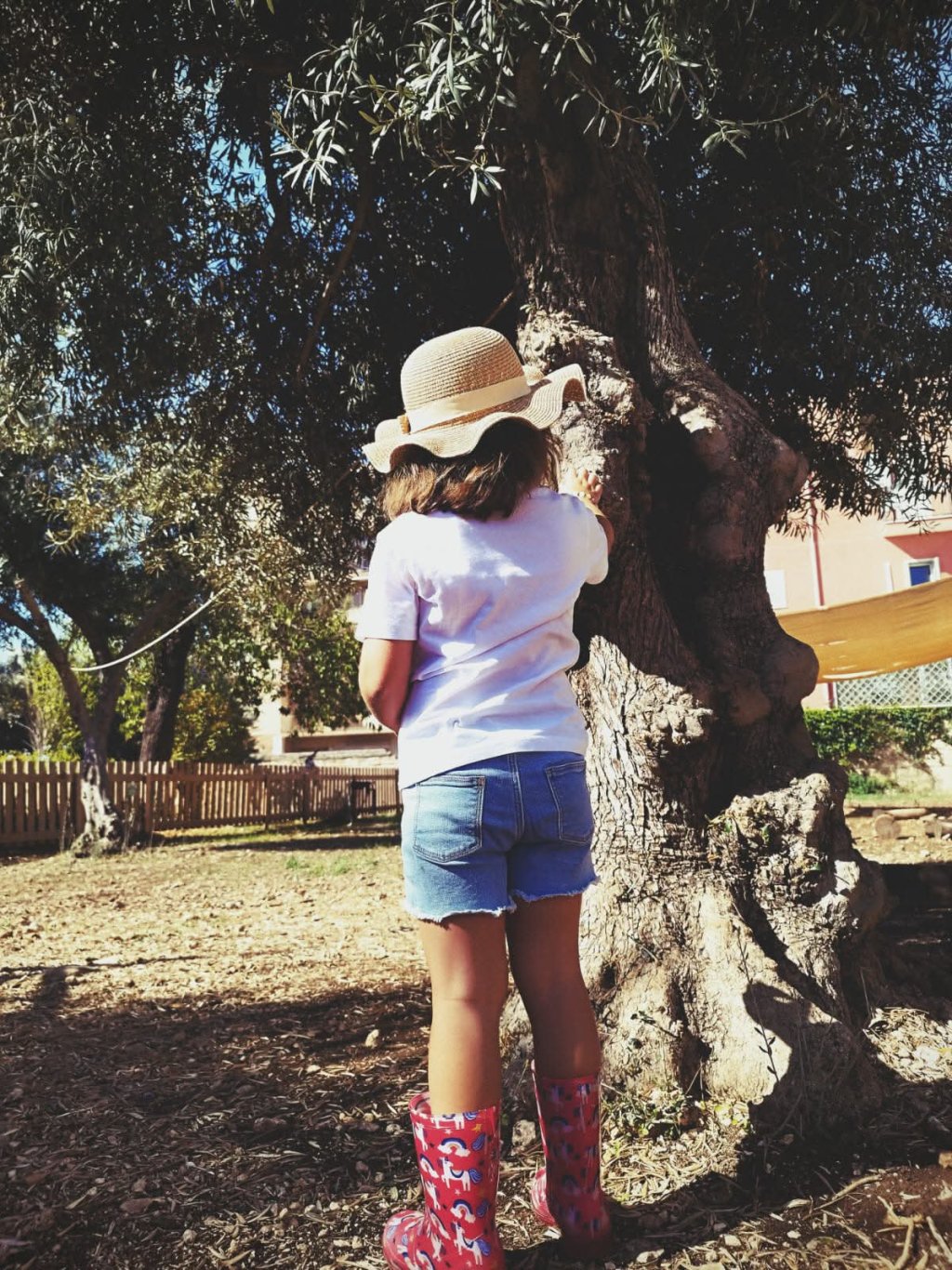 Una bambina indossa cappello, maglietta, short e stivali, si appoggia a un albero