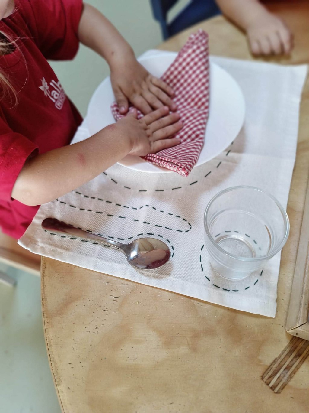 Bambina si asciuga le mani con un panno a quadretti rosso su un tavolo con piatto, bicchiere d'acqua e cucchiaio