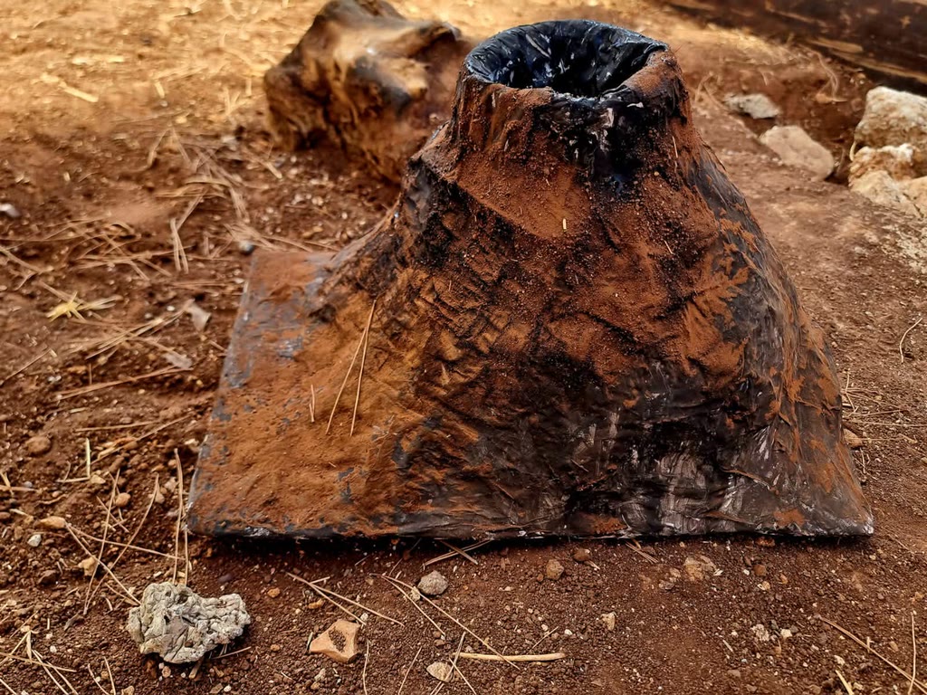 Una sacca di plastica nera coperta di fango è posata su terra bruna con rametti e sassi