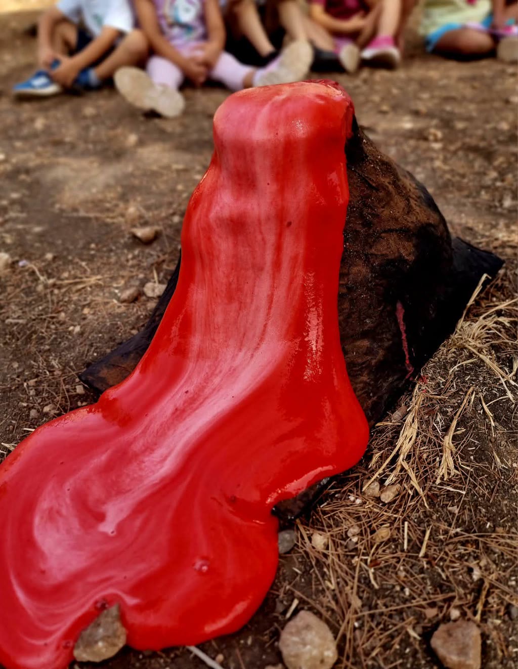 Un vulcano di fango rosso finto è spuntato da un tronco su terreno erboso con bambini seduti in primo piano