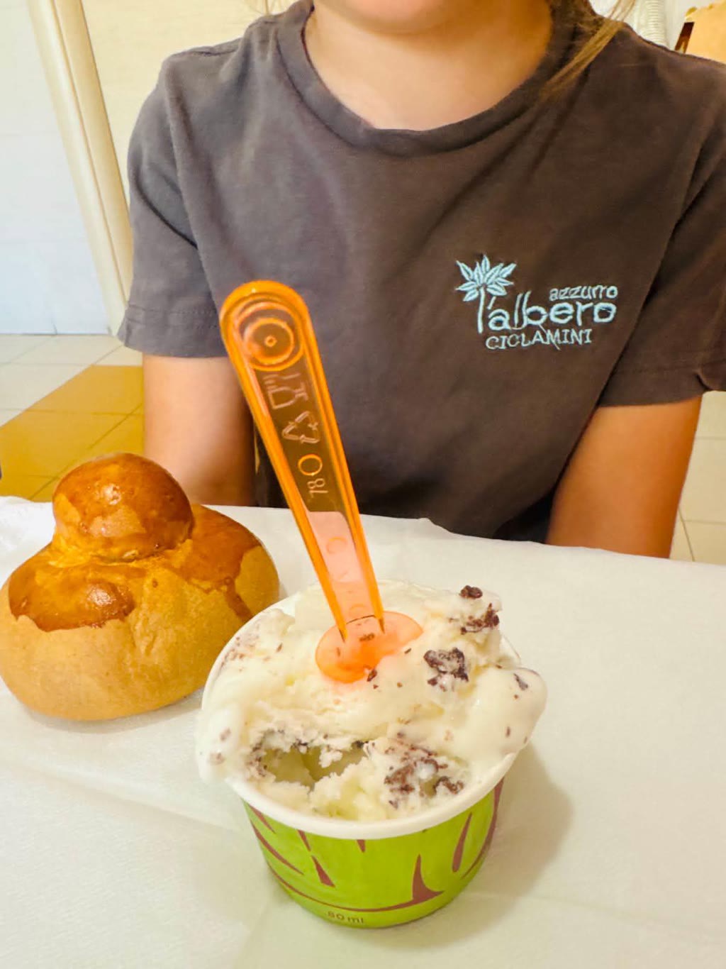 Una persona indossa una maglietta con scritta e tiene un gelato con cialda e un cucchiaio di plastica. Accanto c'è un panino