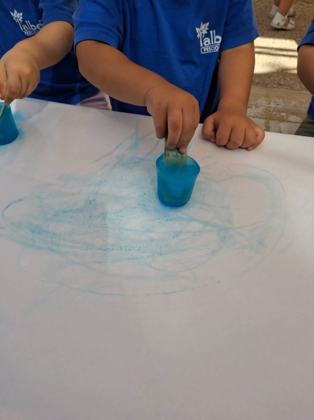 Bambini in maglietta blu fanno scorrere cubetti di ghiaccio colorato su un grande foglio, che si tinge di azzurro.