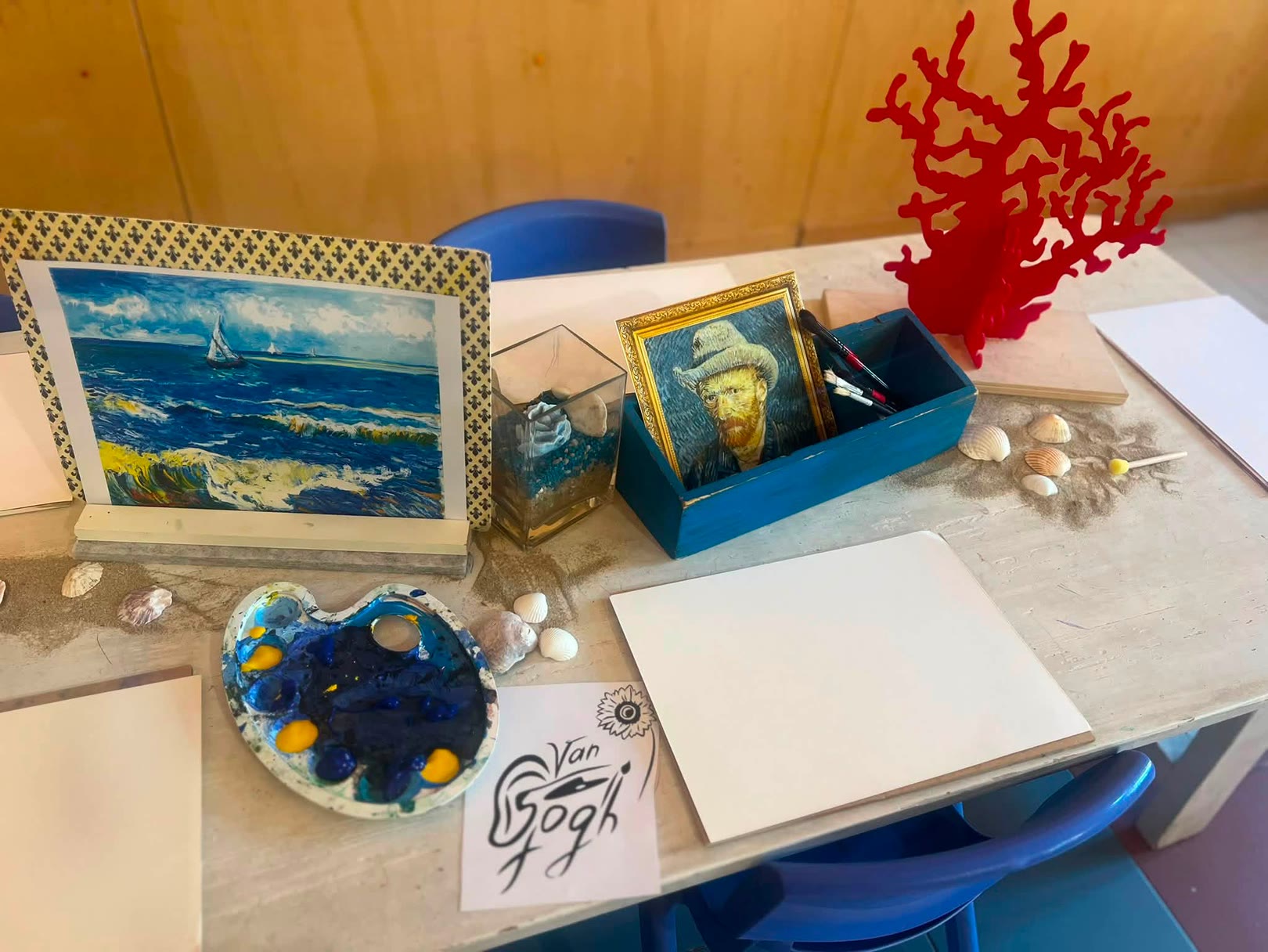 Tavolo con quadro marino, scatola con pennelli, tela, vassoio di colori, conchiglie, carta, e decorazione rossa