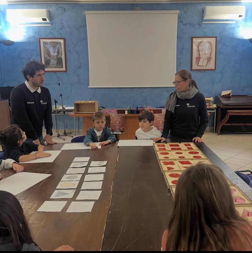 Due adulti e tre bambini si trovano attorno a un tavolo con carte e forme geometriche, in un ambiente scolastico