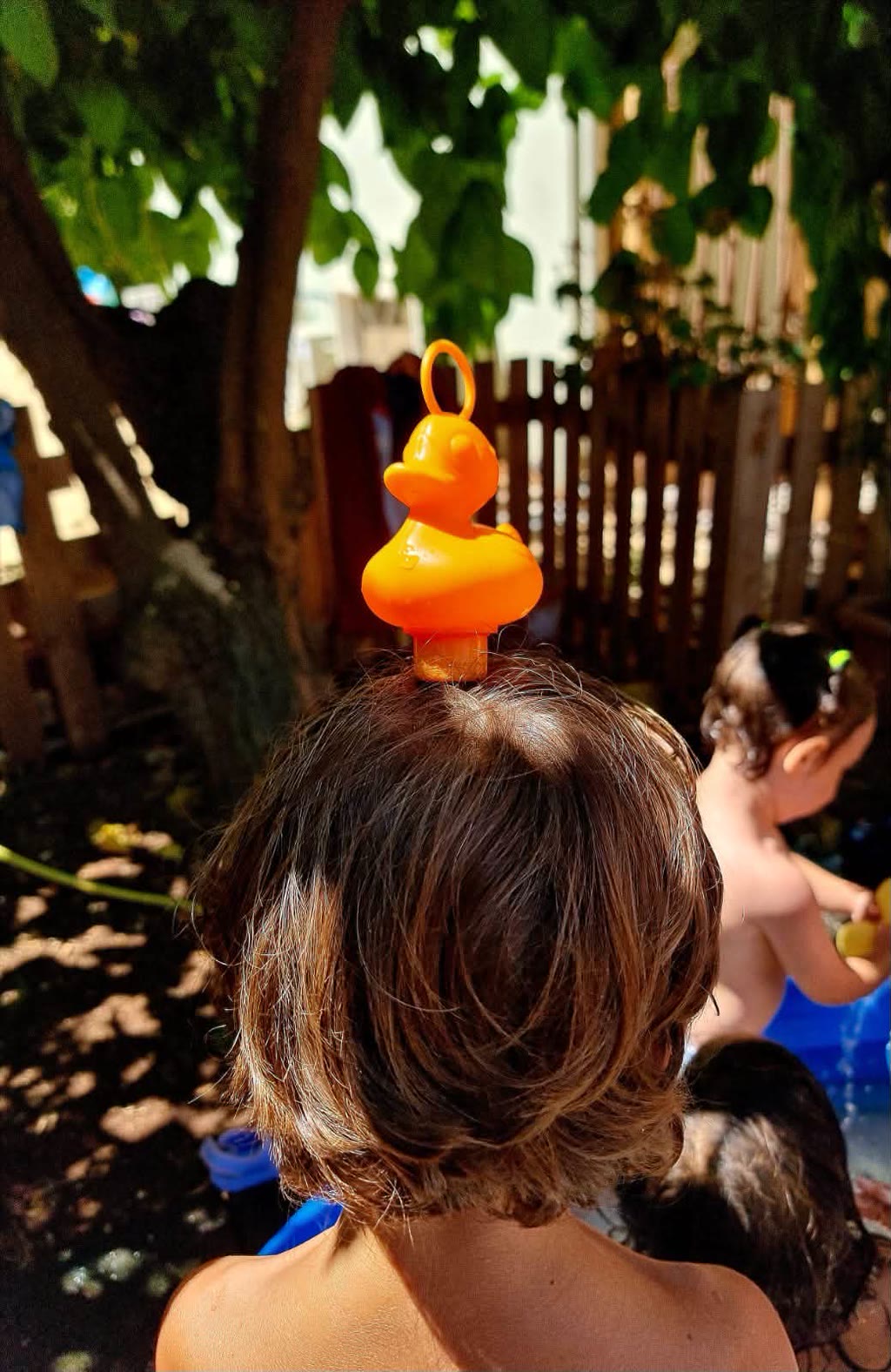 Una bambina con un anello da bagno a forma di gabbiano sulla testa, in un giardino con alberi e recinzione