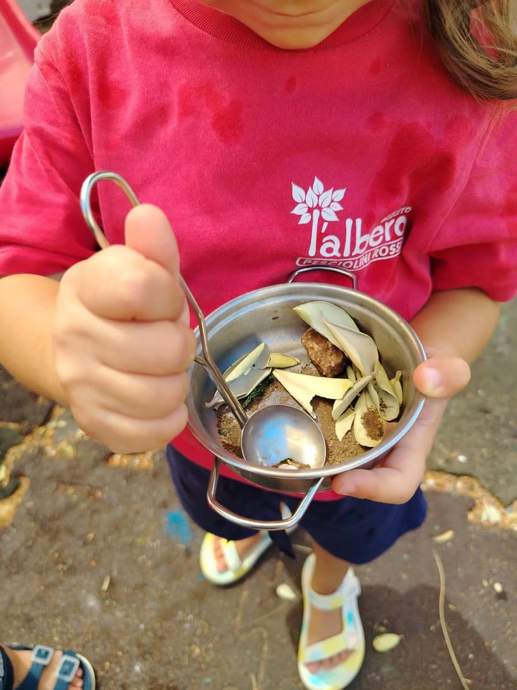 Una bambina in maglietta rossa tiene una ciotola di metallo con una cucchiaio, dentro c'è il rimasugli di cibo