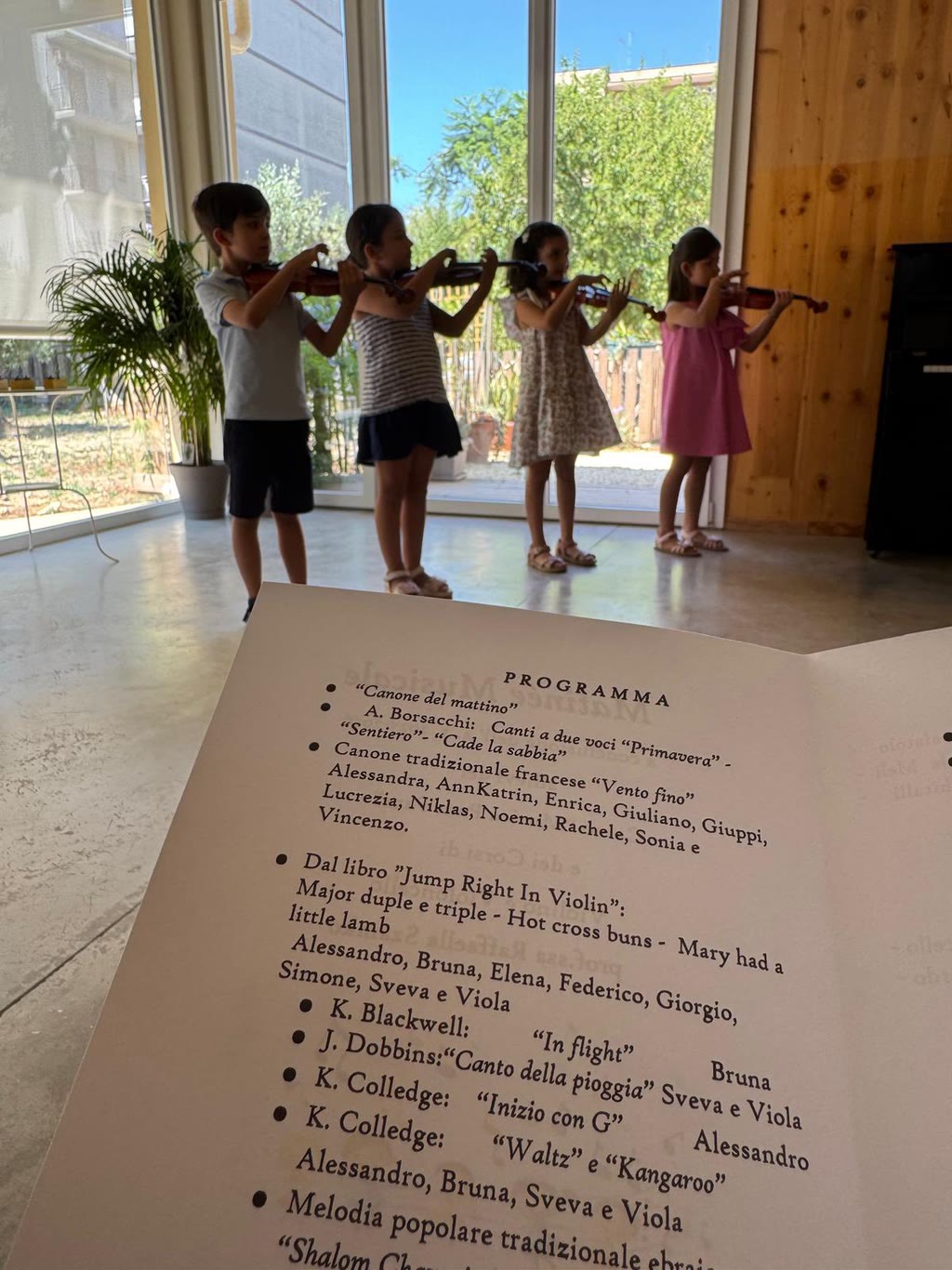 Quattro bambini suonano violini in una stanza con grandi finestre, davanti a un braccio musicale