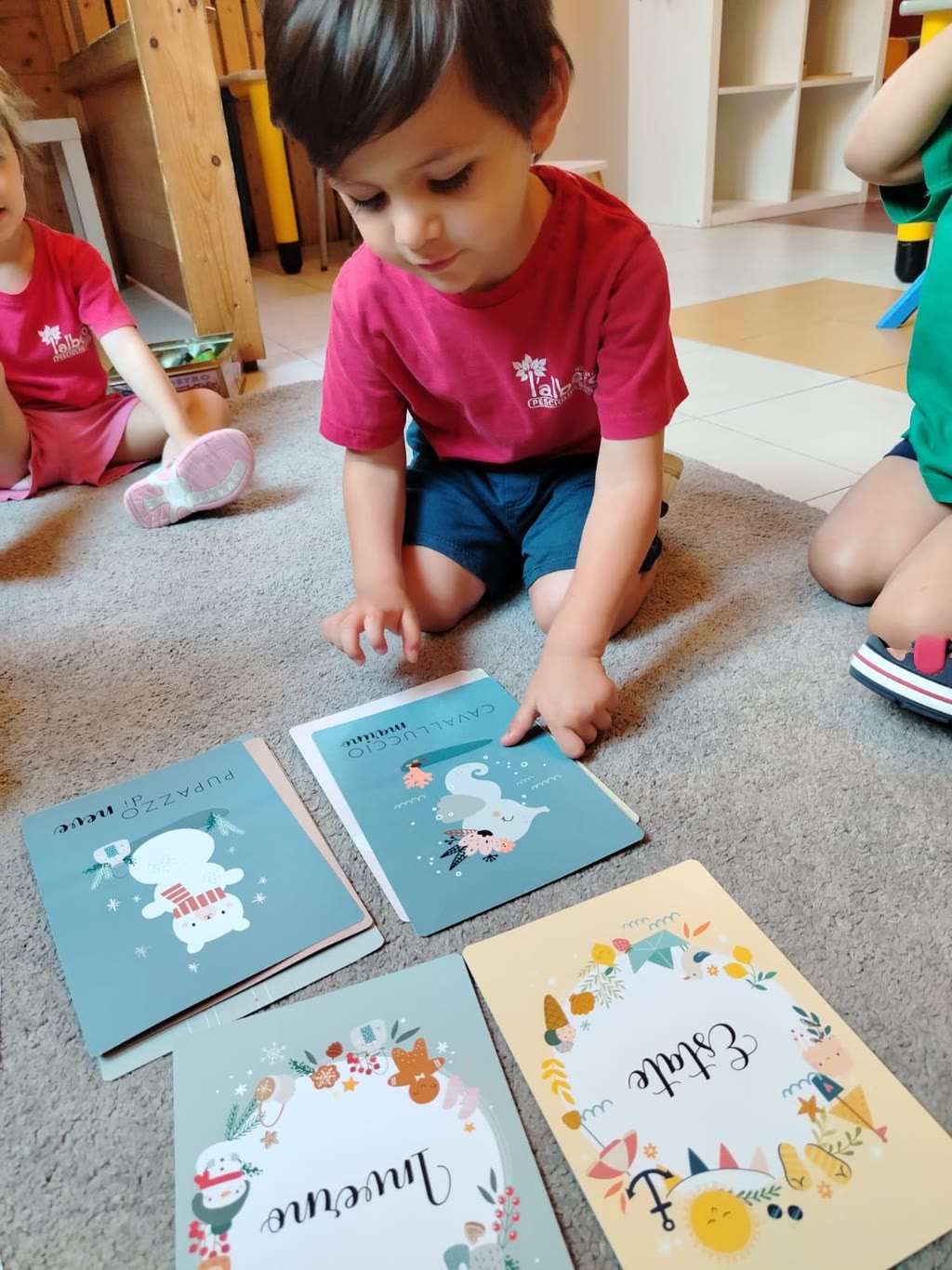 Un bambino in maglietta rossa si siede sul pavimento, puntando su carte colorate con illustrazioni e scritte