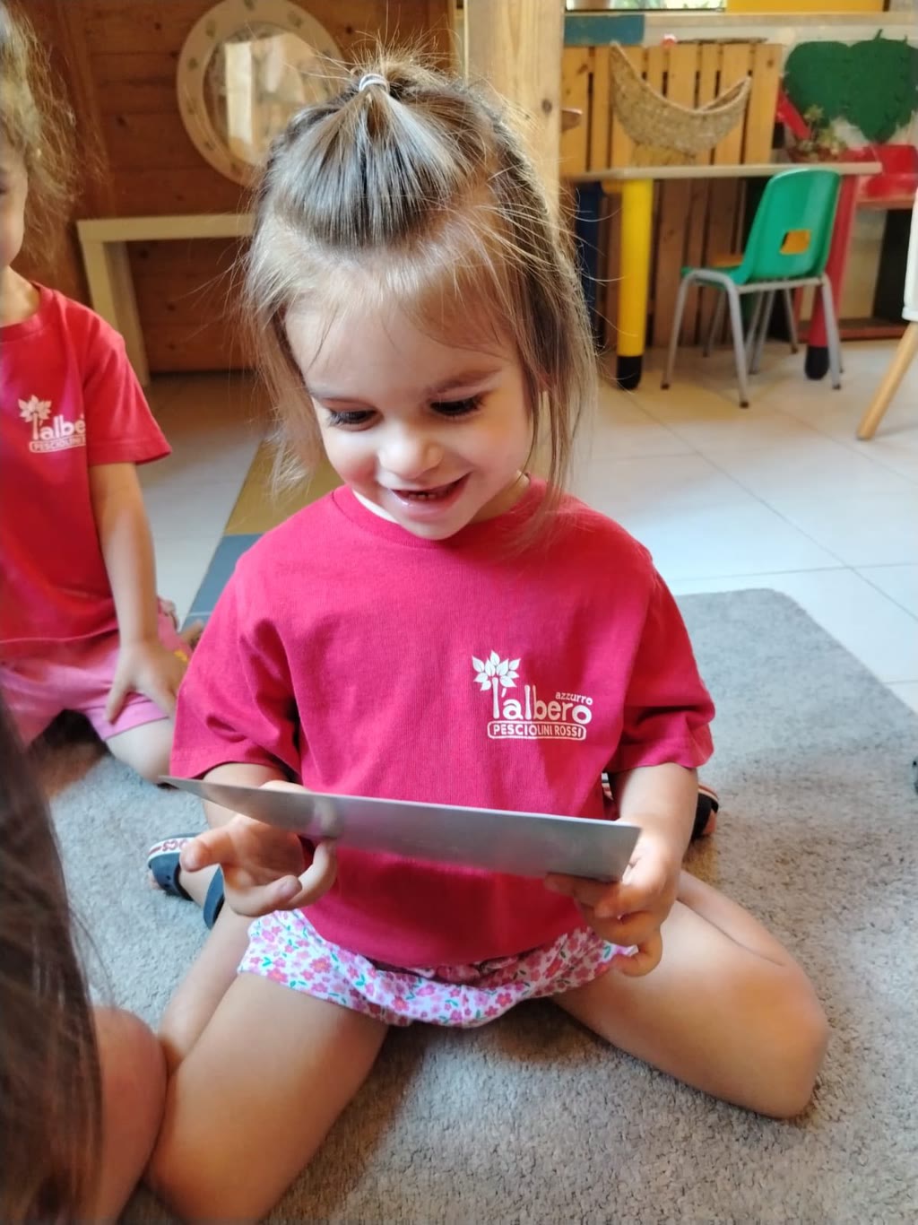 Bambina in maglietta rossa con scritta, seduta sul pavimento, tiene in mano un oggetto lungo