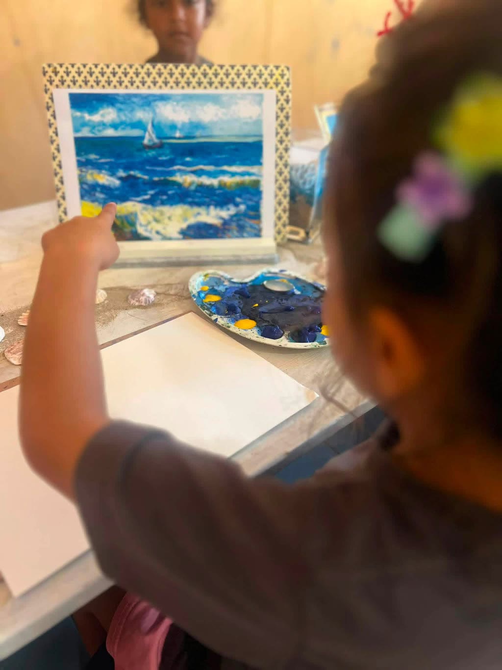 Una bambina osserva un dipinto marino mentre un altro bambino tiene in mano un quadro con una barca a vela