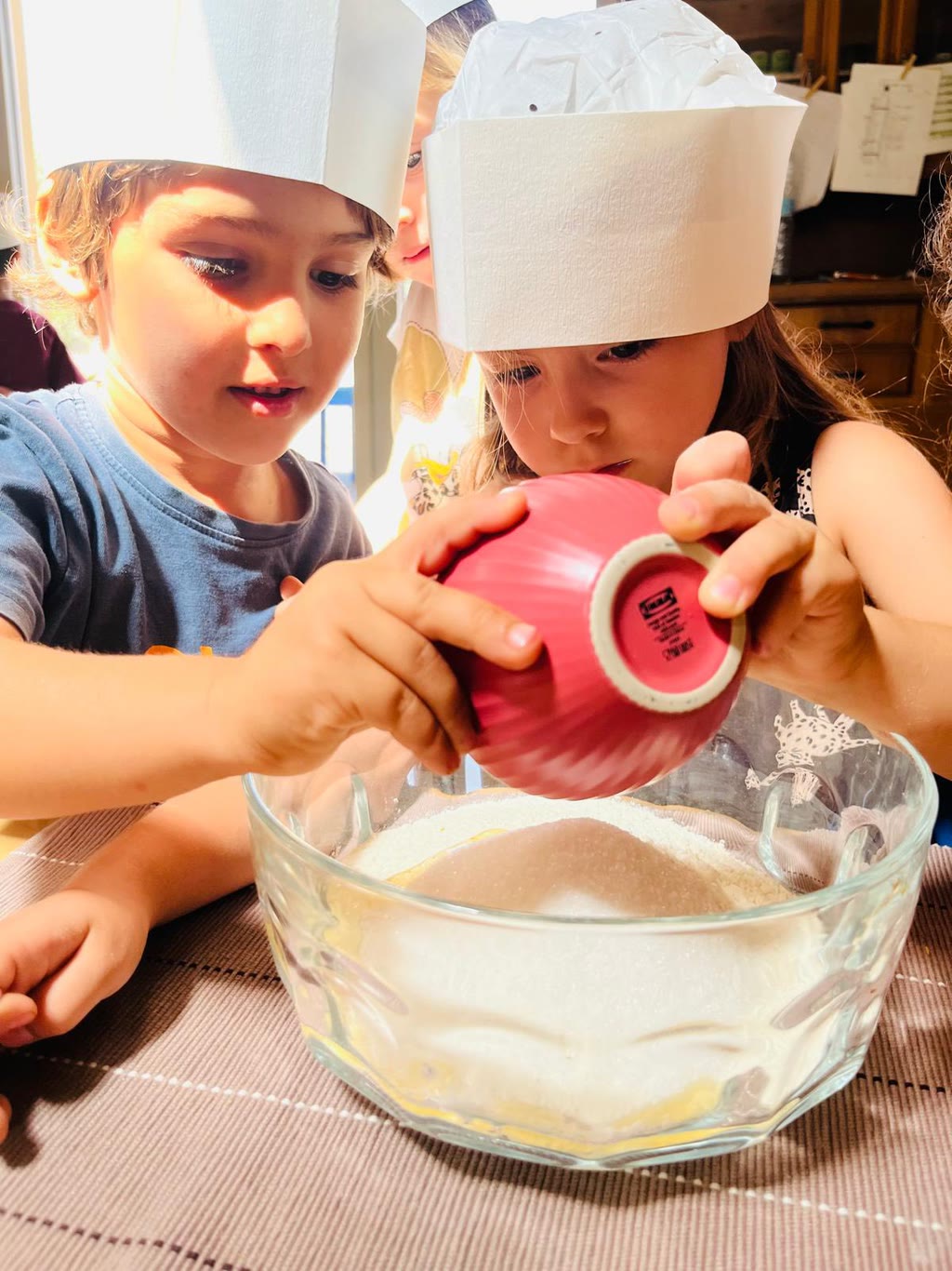 Due bambini indossano cappelli da cuoco e versano farina in una grande ciotola di vetro