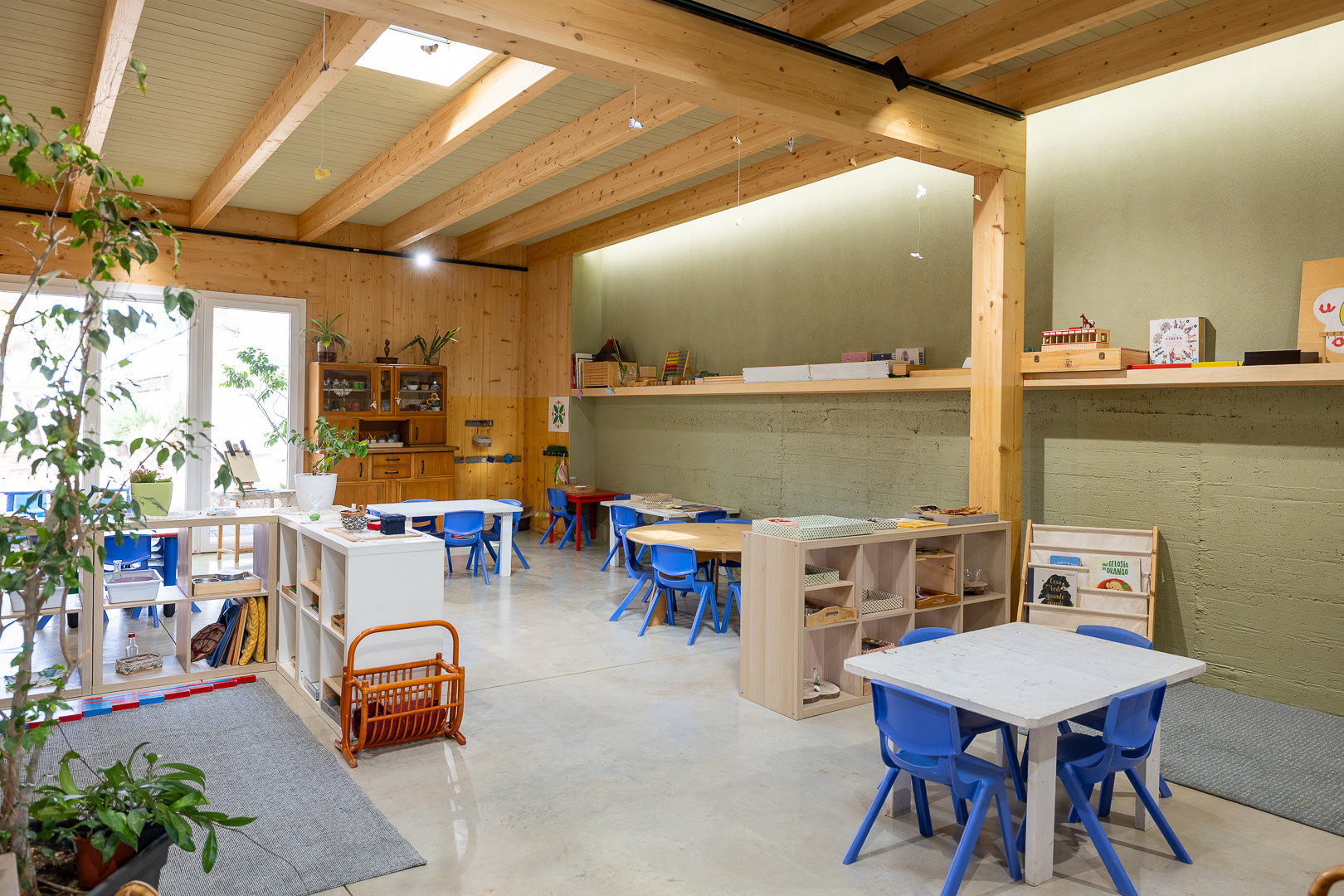 Aula con piante alte, scaffali aperti e tavoli rotondi sotto il soffitto di travi.