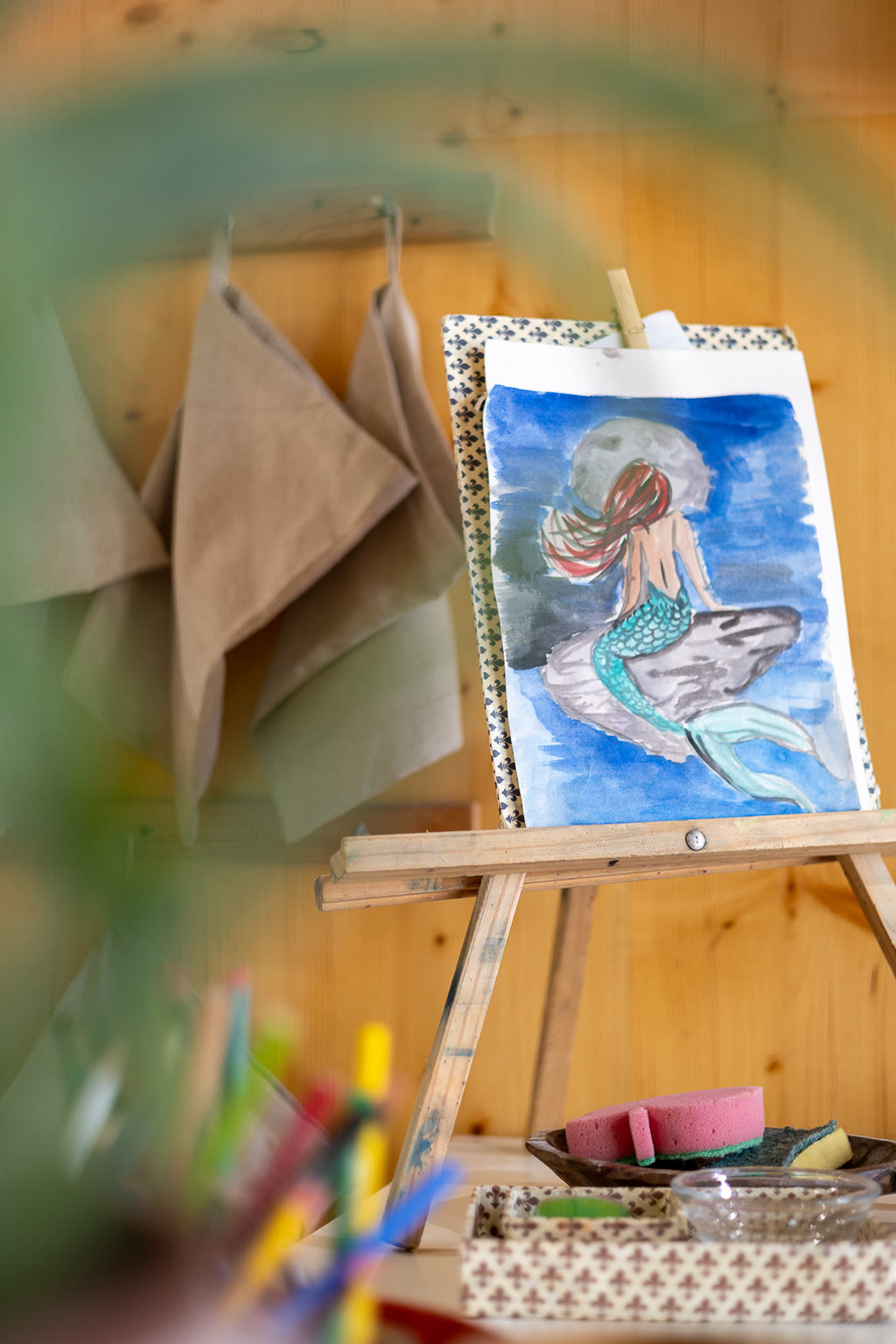 Cavalletto di legno con un acquerello di una sirena, accanto ai grembiuli appesi.