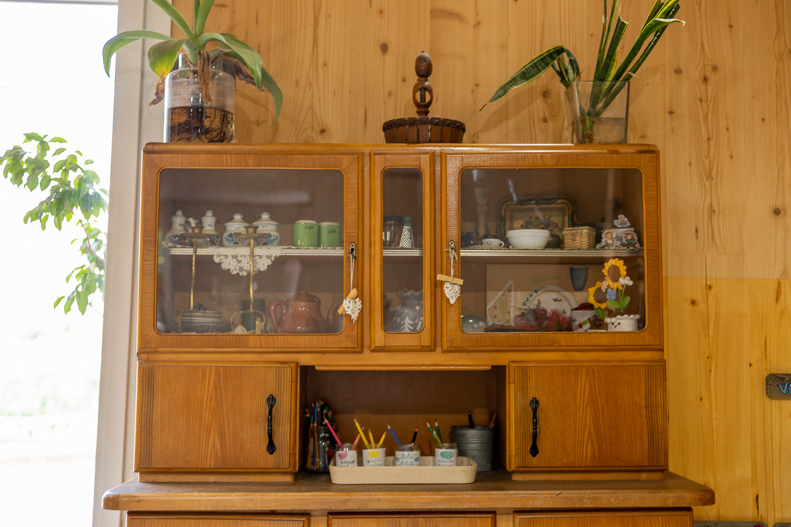 Credenza di legno con ante a vetri, stoviglie vere e piante sopra il ripiano.