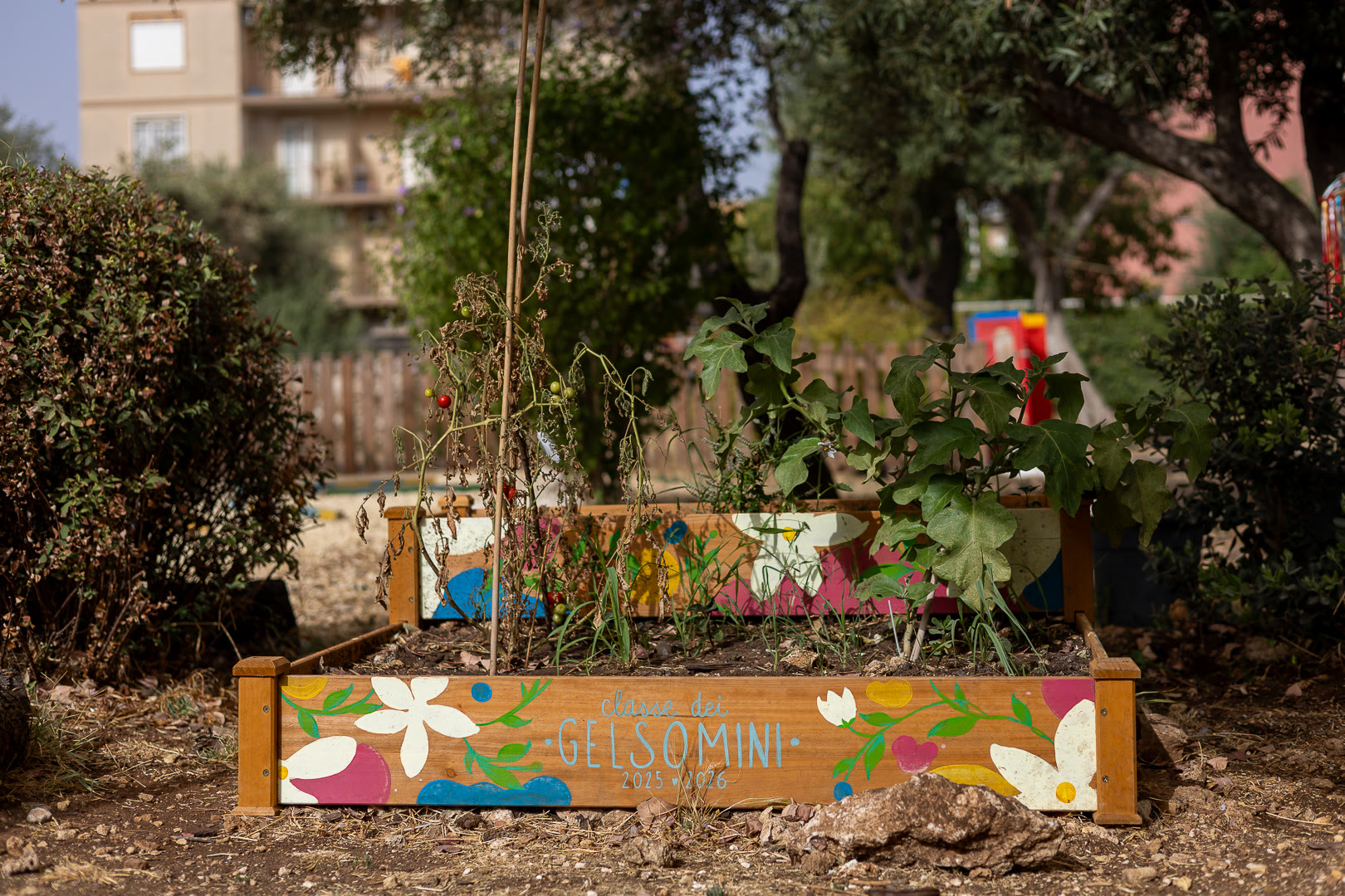 Fioriera di legno dipinta a fiori con la scritta Gelsomini, piantata a verde.