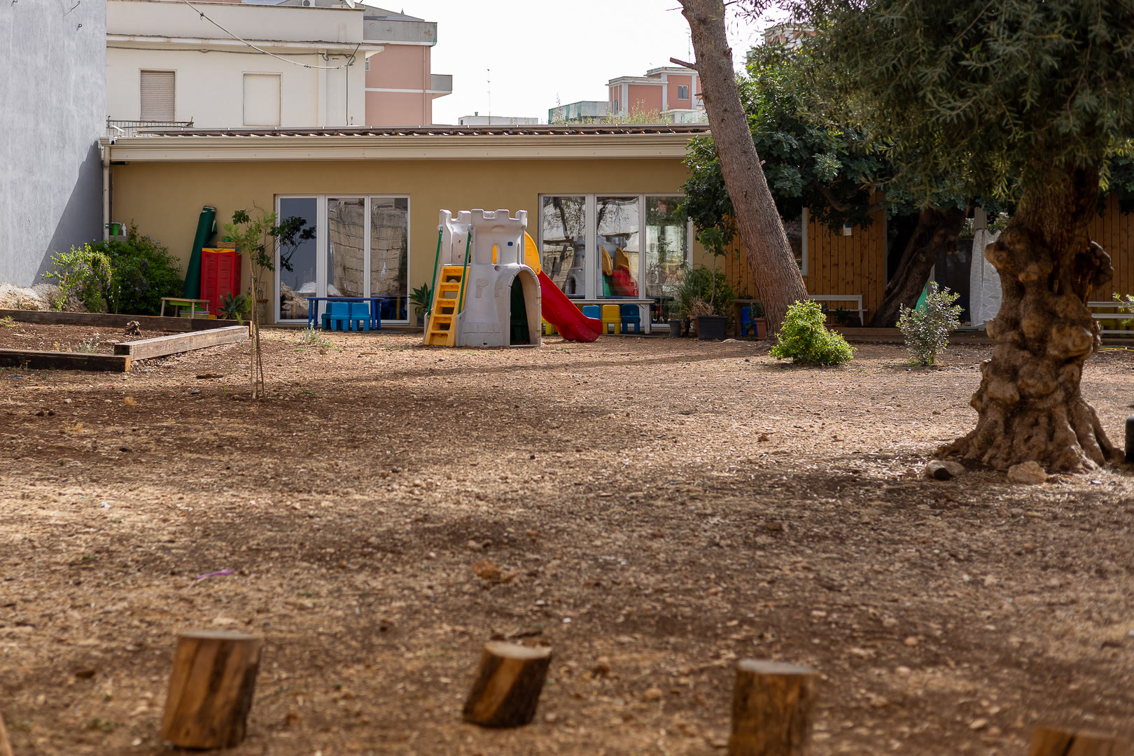 Cortile in terra battuta con lo scivolo, gli ulivi e i ceppi di legno usati come sedute.