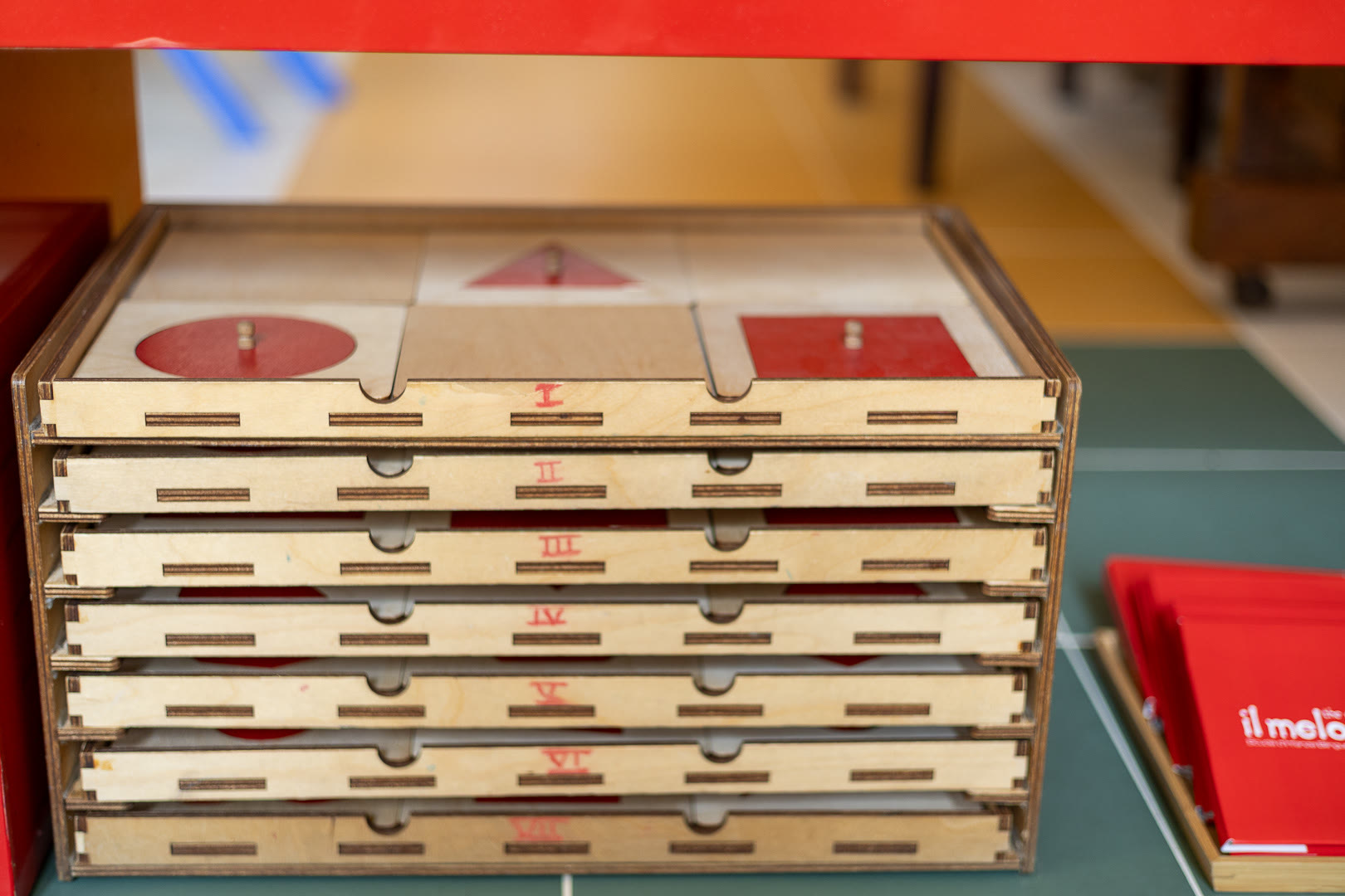 Cassettiera degli incastri geometrici: i cassetti numerati in cifre romane e le figure rosse — cerchio, triangolo, quadrato — nel ripiano in alto.