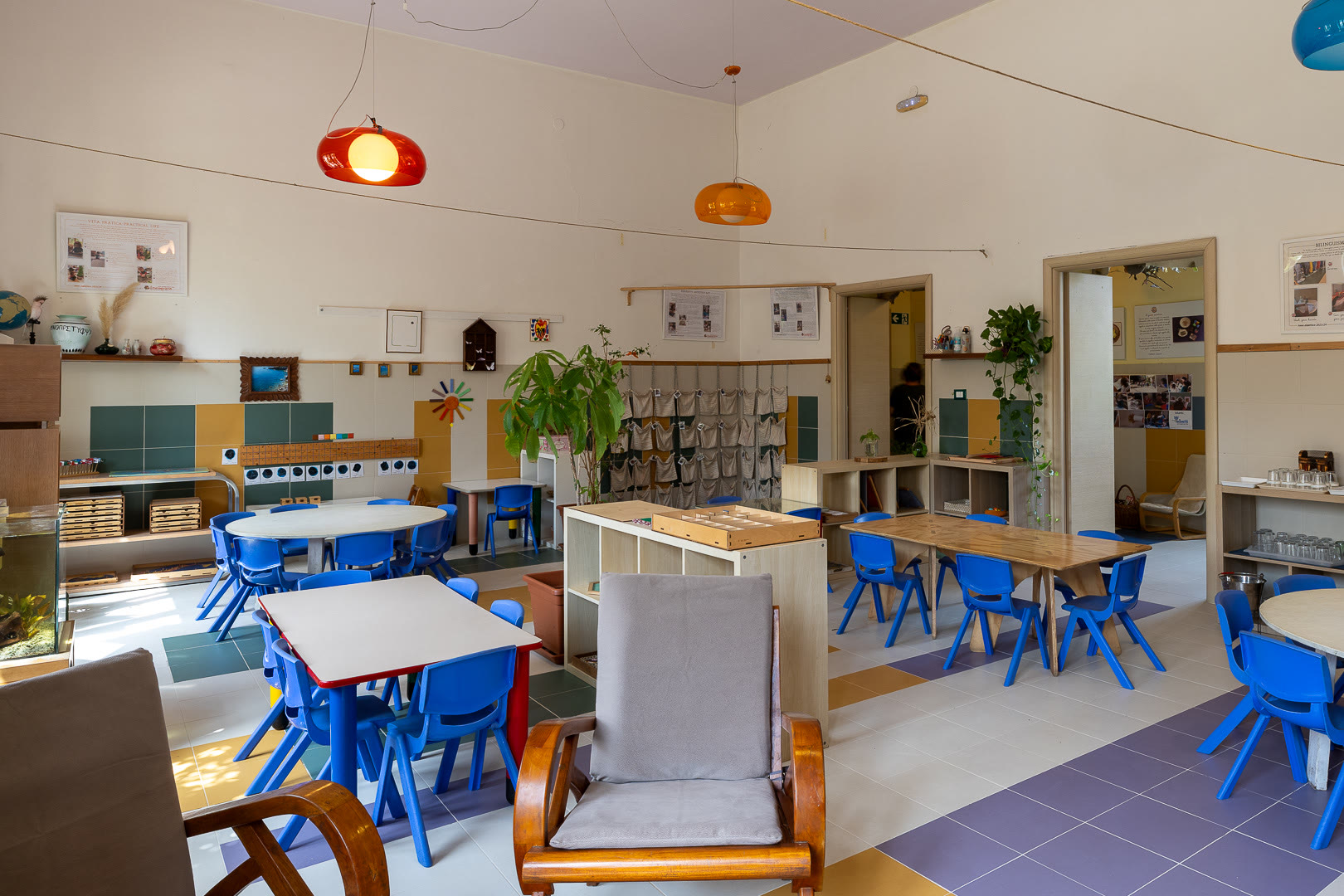 Ambiente della Casa dei Bambini con il soffitto alto, le lampade arancioni e i tavoli di lavoro.