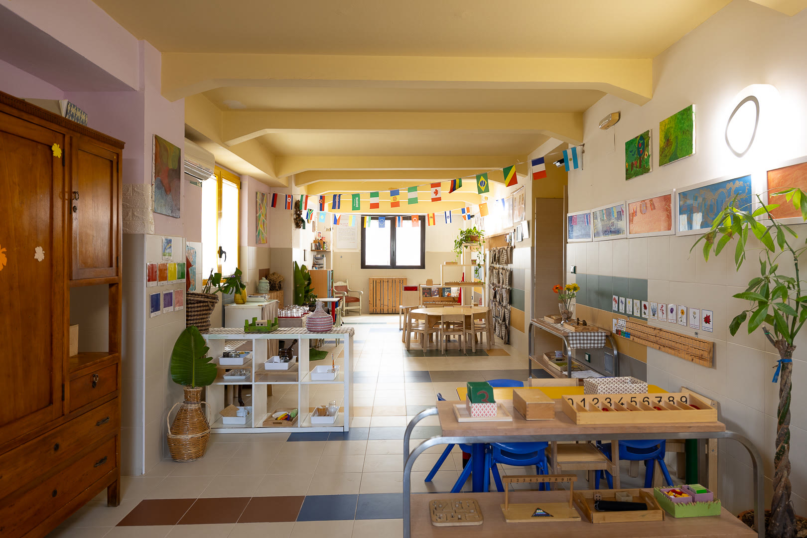 Aula della Casa dei Bambini: scaffali bassi, tavoli in legno e piante alle finestre.