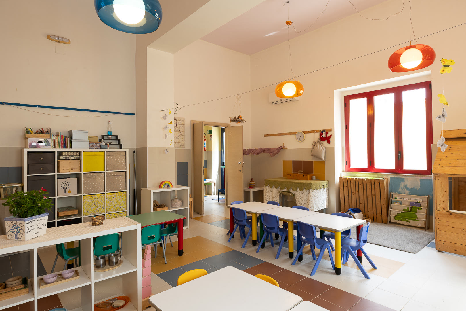 Aula con scaffali aperti, tavoli colorati e la finestra dagli infissi rossi.