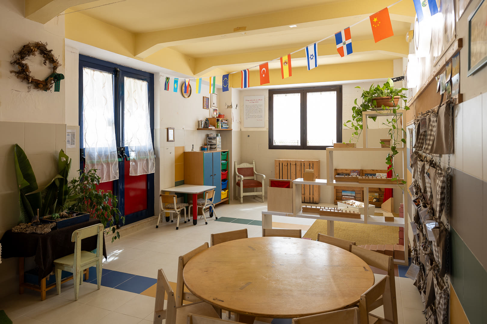 Aula con il tavolo rotondo in legno, le sedie dei bambini e il materiale sugli scaffali.