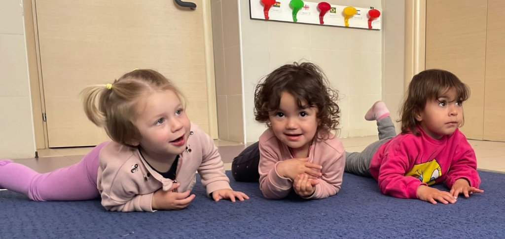 Tre bambine sdraiate a pancia in giù sul tappeto, in ascolto.