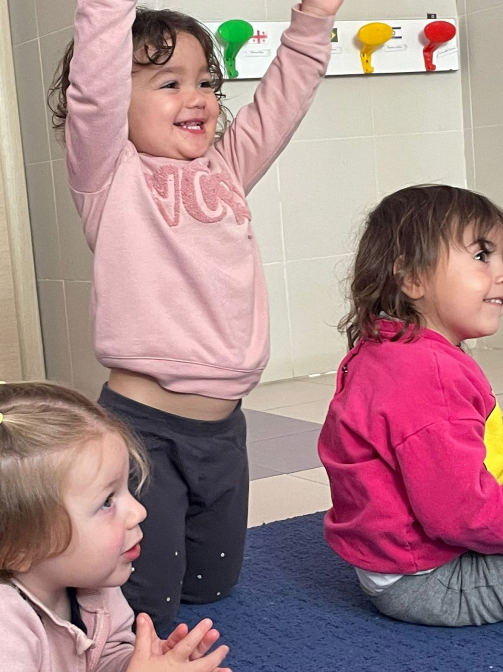 Una bambina in ginocchio sul tappeto alza le braccia e ride, accanto a due bambine sedute.