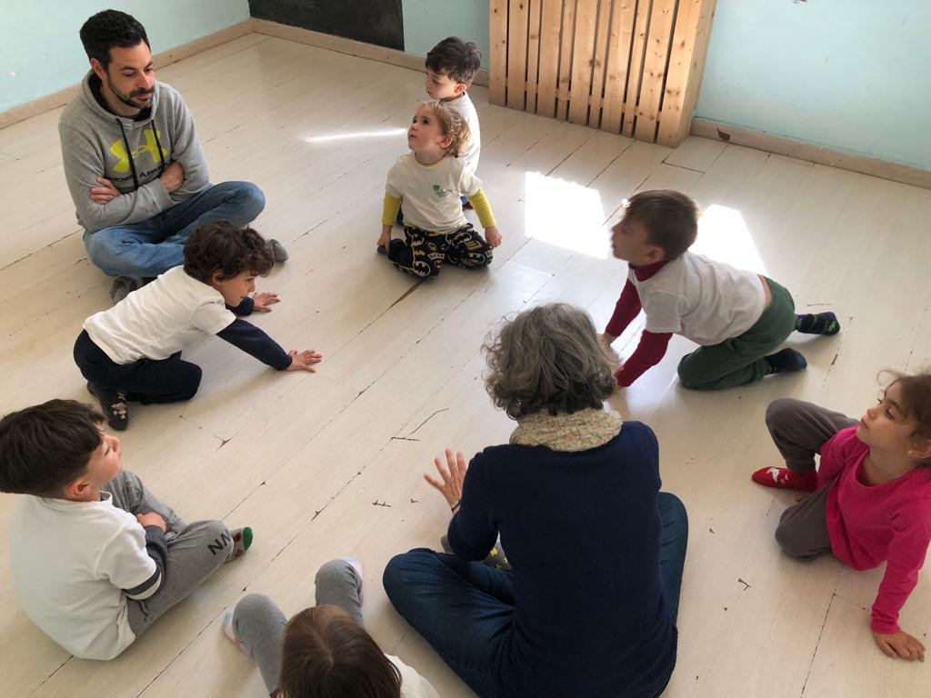 Bambini seduti in cerchio sul pavimento con due adulti, durante un gioco.