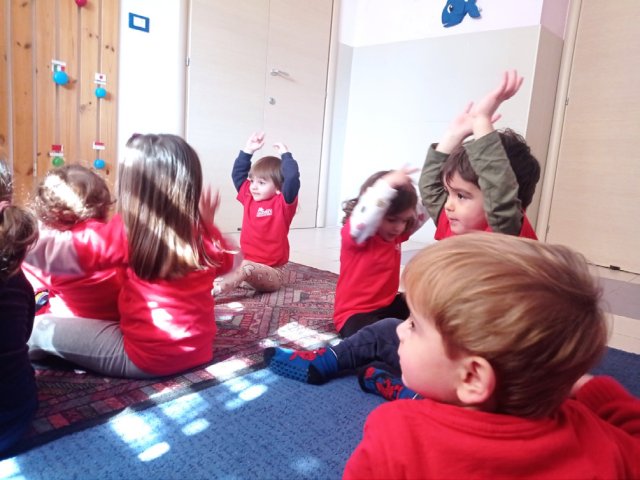 Bambini con la maglia rossa, seduti sul tappeto, alzano le mani sopra la testa.